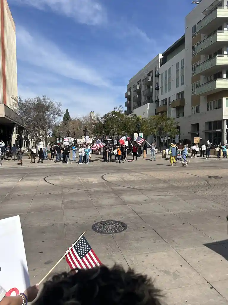 A wide-angle photo of a public protest or rally on a sunny day, with many people holding American flags and signs in a city…