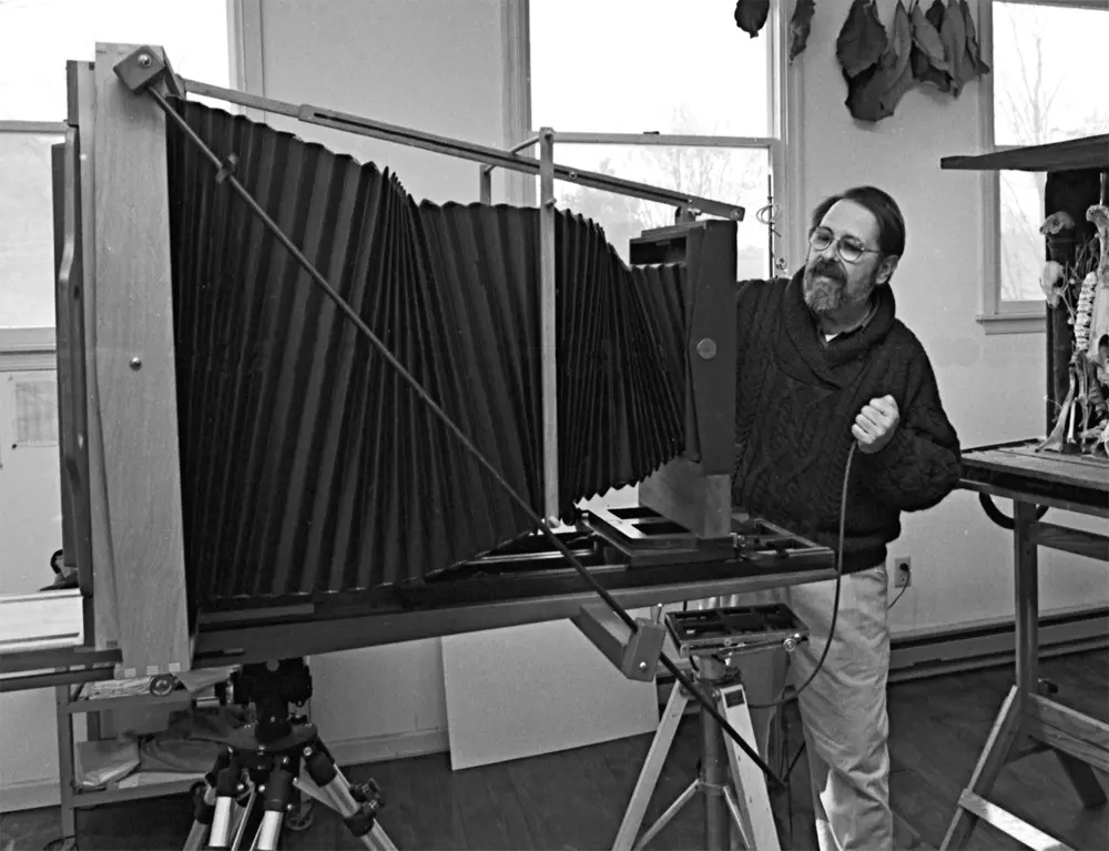 A black-and-white photo shows a man with glasses and a beard standing beside a large-format view camera with an extended…