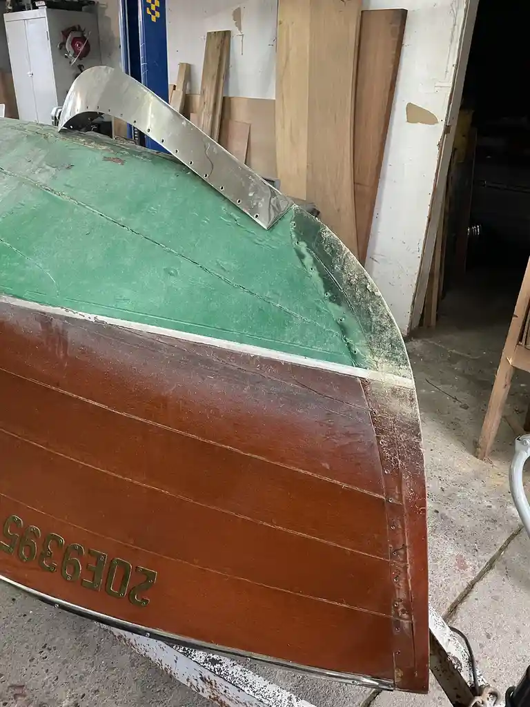 A close-up of a wooden boat's bow in a workshop, showing a newly installed shiny metal bow rail bolted to the green and…