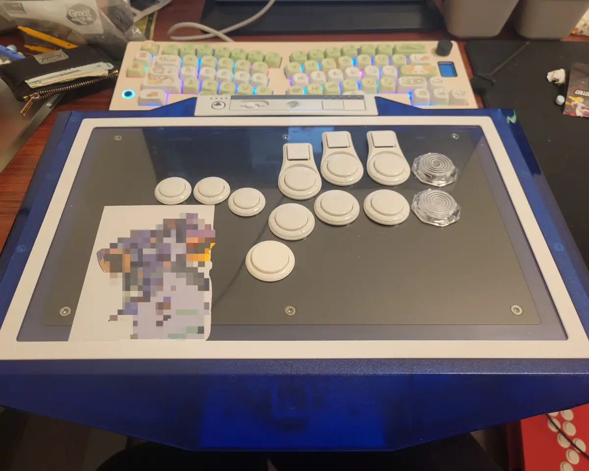 A custom-built arcade stick with white buttons and clear analog sticks sits on a desk, featuring a pixelated image of a…