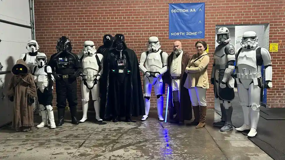 A group of people dressed as Star Wars characters, including Darth Vader, Stormtroopers, and Jedi, pose for a photo in front…