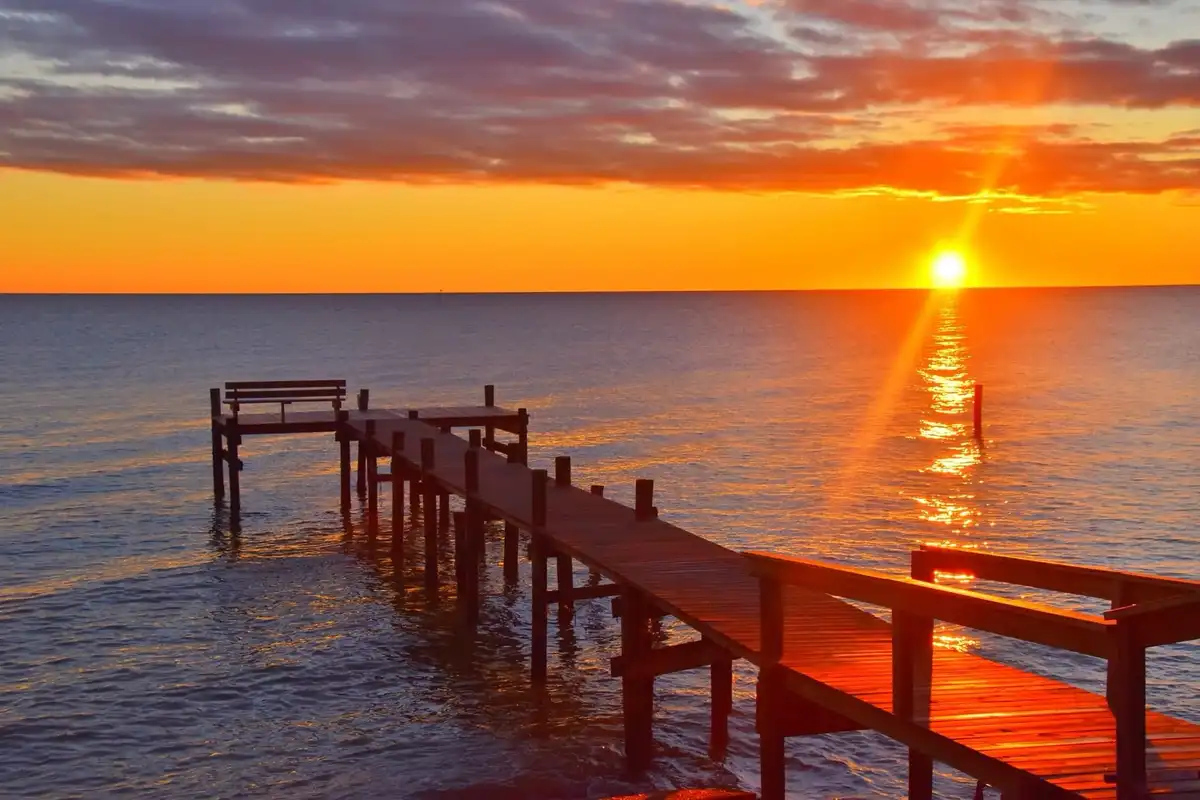 A serene sunset over the ocean with a wooden pier extending into the water, featuring a bench at its end. The sun is low on…