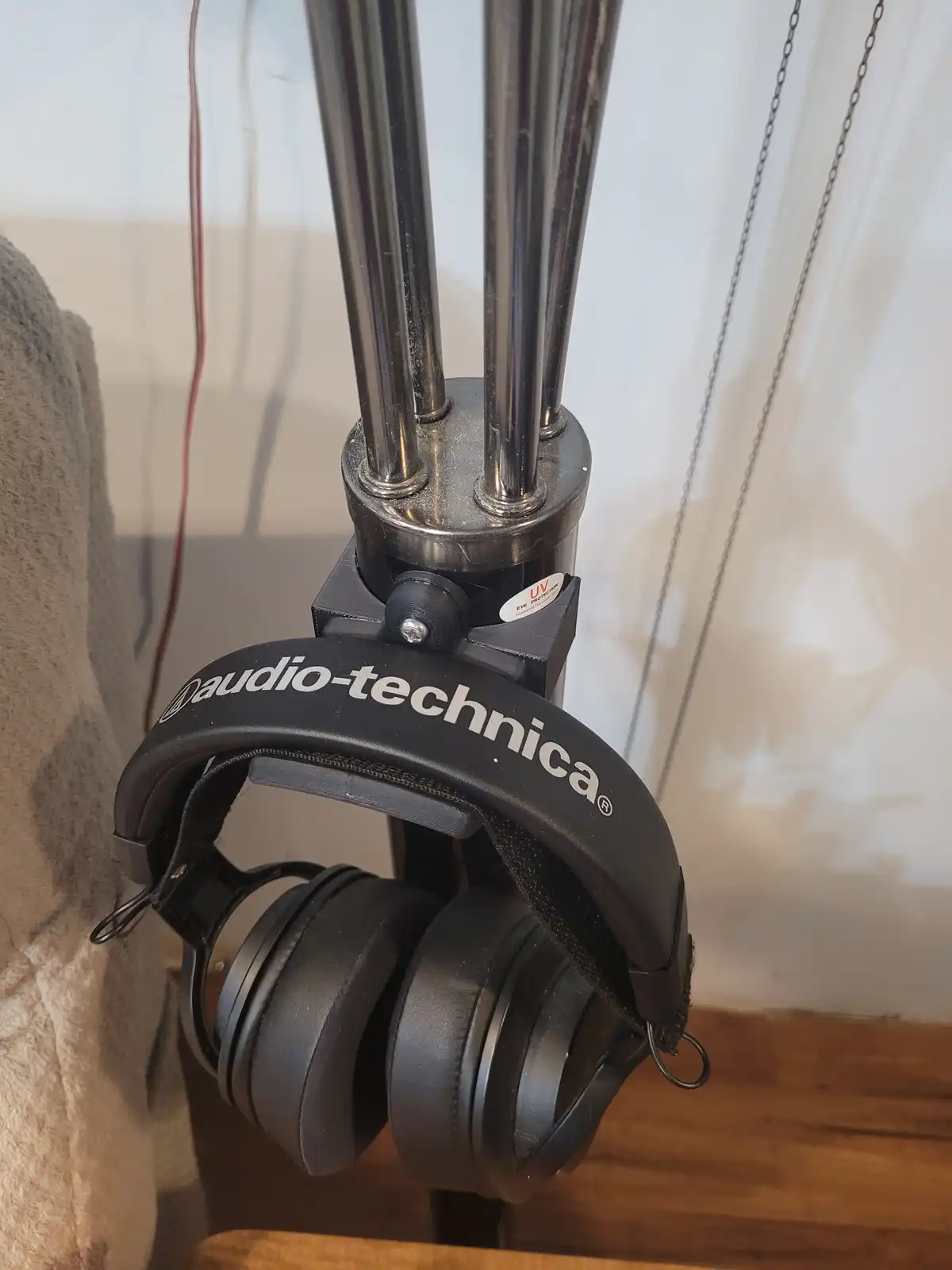 A pair of black Audio-Technica over-ear headphones hangs from a metal tripod stand, with a small white sticker reading 'UV…