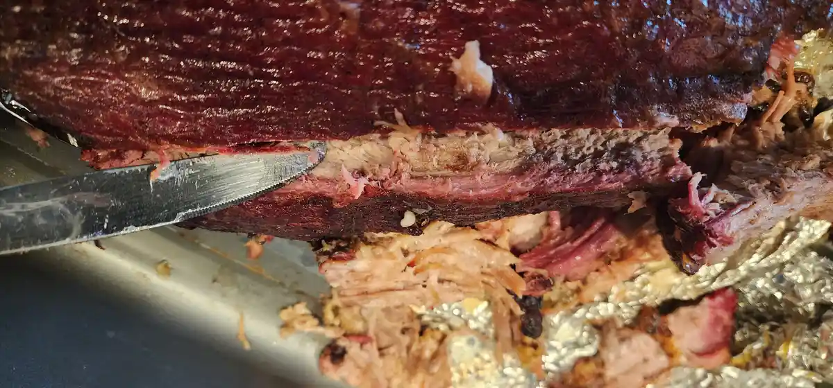 A close-up photo shows a knife slicing into a large, darkly smoked brisket with tender, shredded meat falling away. The meat…