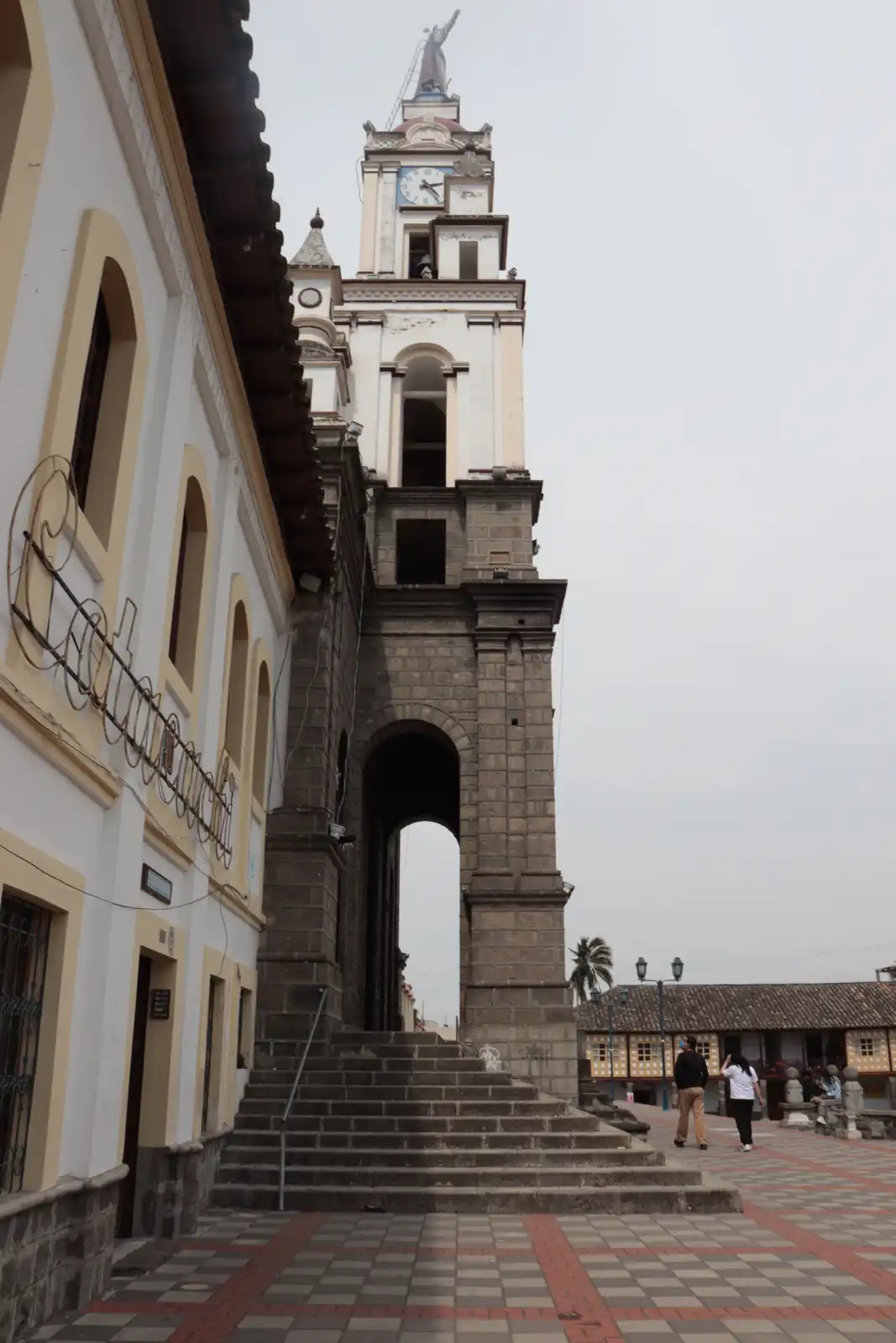 A low-angle photo of a historic stone church tower with a clock and statue, flanked by colonial-style buildings and a tiled…