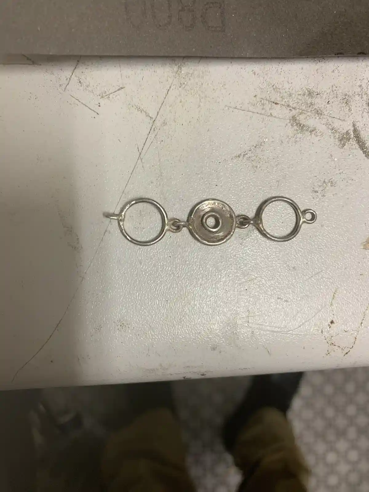 A close-up photo shows a small, silver-colored metal chain with two circular loops connected by a central disc clasp…