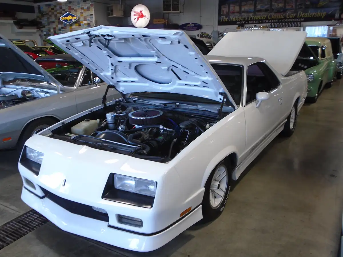 A white classic car with its hood open, revealing the engine, is parked in a classic car dealership. The car's rear hatch is…