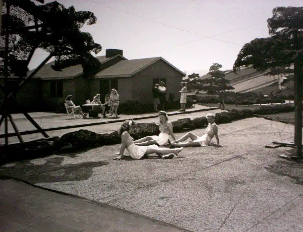 A vintage black-and-white photo shows three women lounging on a paved patio, appearing relaxed and sunbathing. In the…