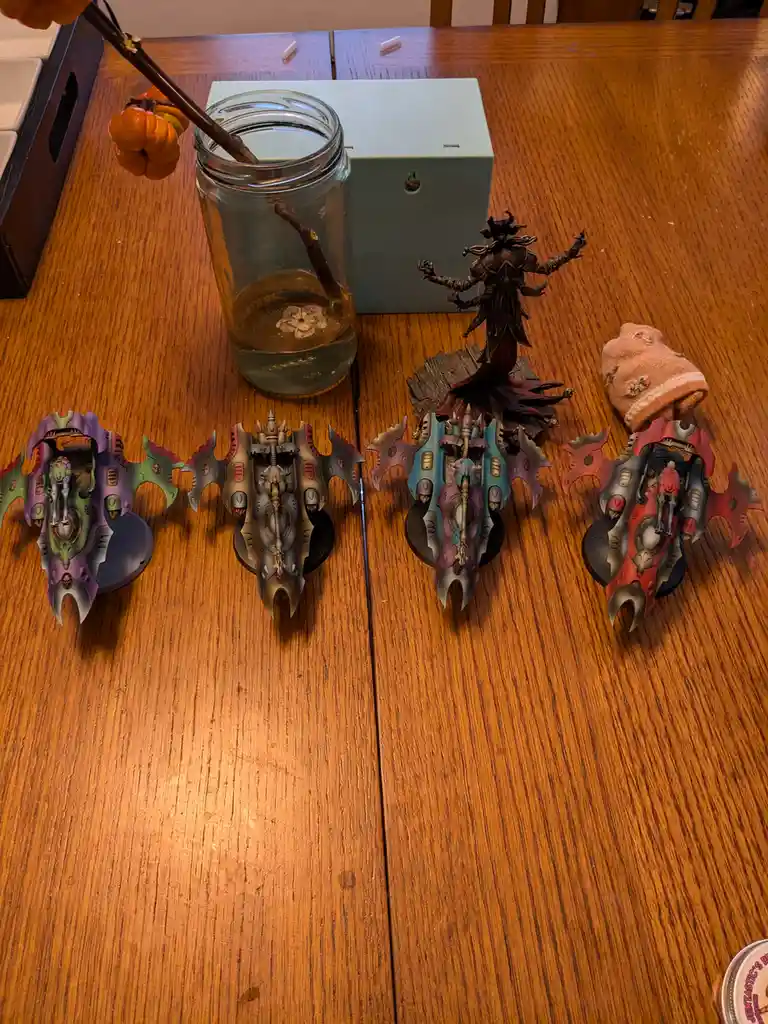 Four painted Warhammer 40k miniatures are arranged on a wooden table, with a large multi-armed dark figure standing behind…