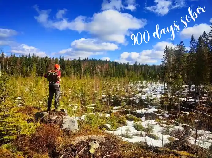 A person wearing an orange beanie, red shirt, and green boots stands on a rock overlooking a forest with patches of snow…