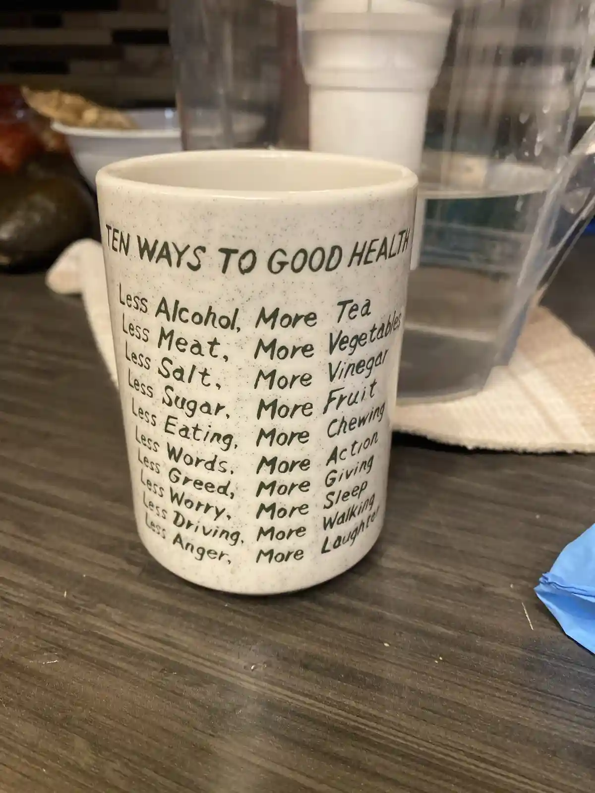 A white speckled ceramic mug sits on a dark wooden countertop, printed with 'TEN WAYS TO GOOD HEALTH' and a list of paired…