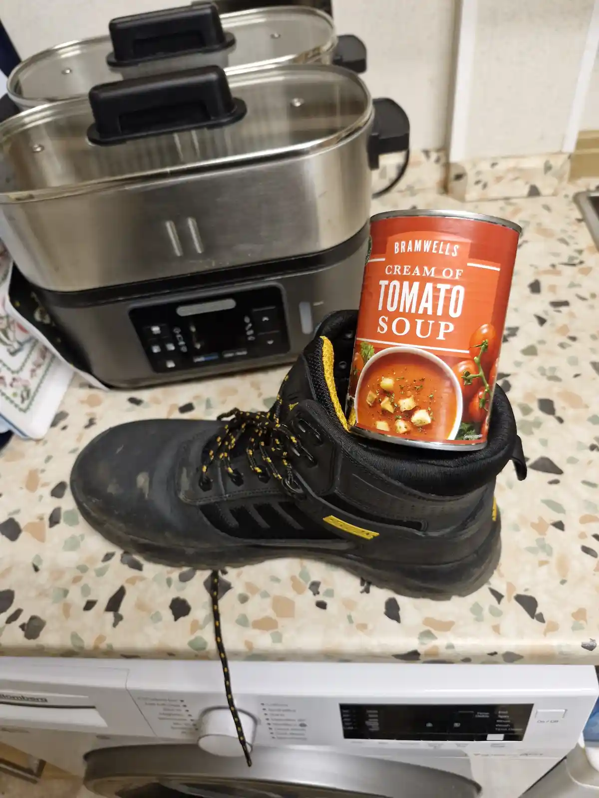 A can of Bramwells Cream of Tomato Soup is stuck upright inside the opening of a black hiking boot, which sits on a speckled…