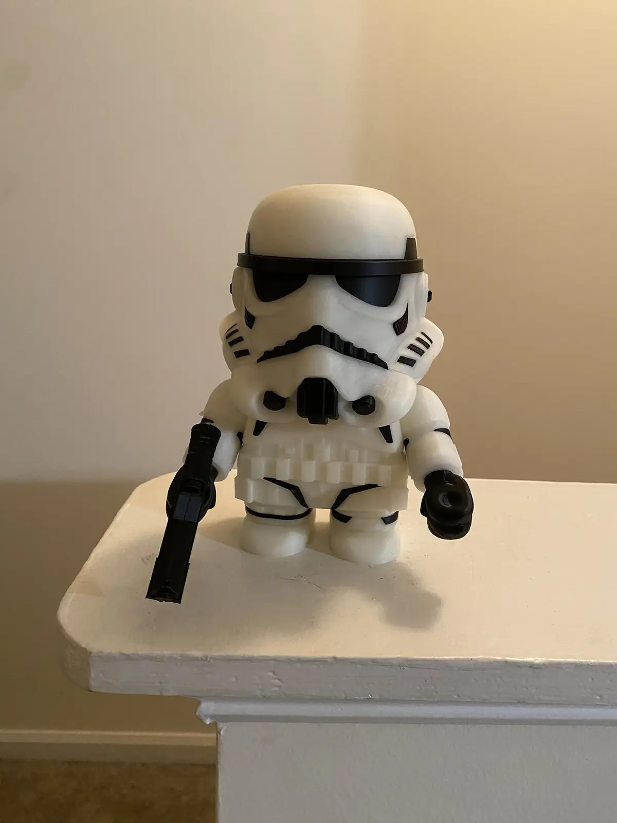 A small, white 3D-printed Stormtrooper figurine from Star Wars stands on a white mantelpiece, holding a black blaster pistol…