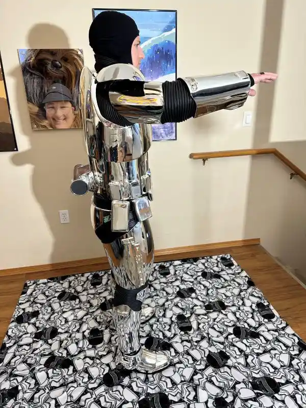 A person wearing a full-body, shiny chrome robot costume with black fabric accents stands indoors, arms outstretched, on a…
