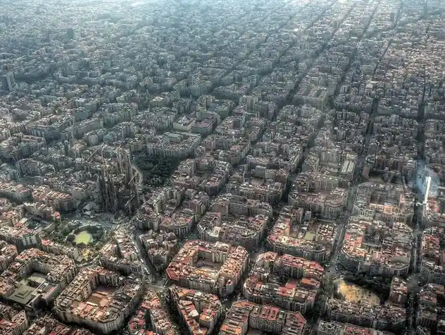 An aerial view of a dense urban grid with a prominent, uniquely shaped cathedral under construction near the center-left…