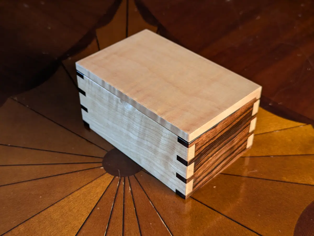 A small, handmade wooden box with a light-colored top and dark-stained sides, featuring visible dovetail joints, sits on a…