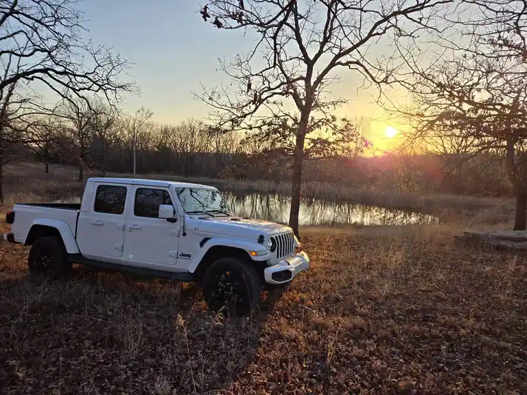 A white Jeep Gladiator pickup truck is parked in a field of dry grass near a body of water, with bare trees silhouetted…