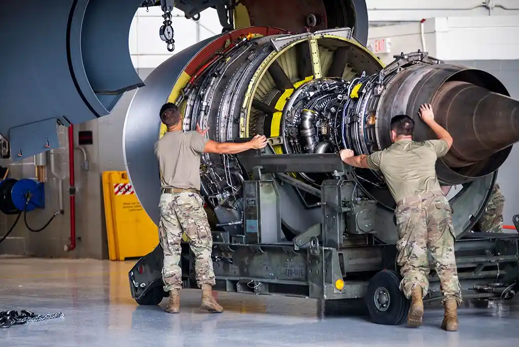 Two men in military camouflage uniforms are pushing a massive jet engine mounted on a wheeled cart inside an aircraft…
