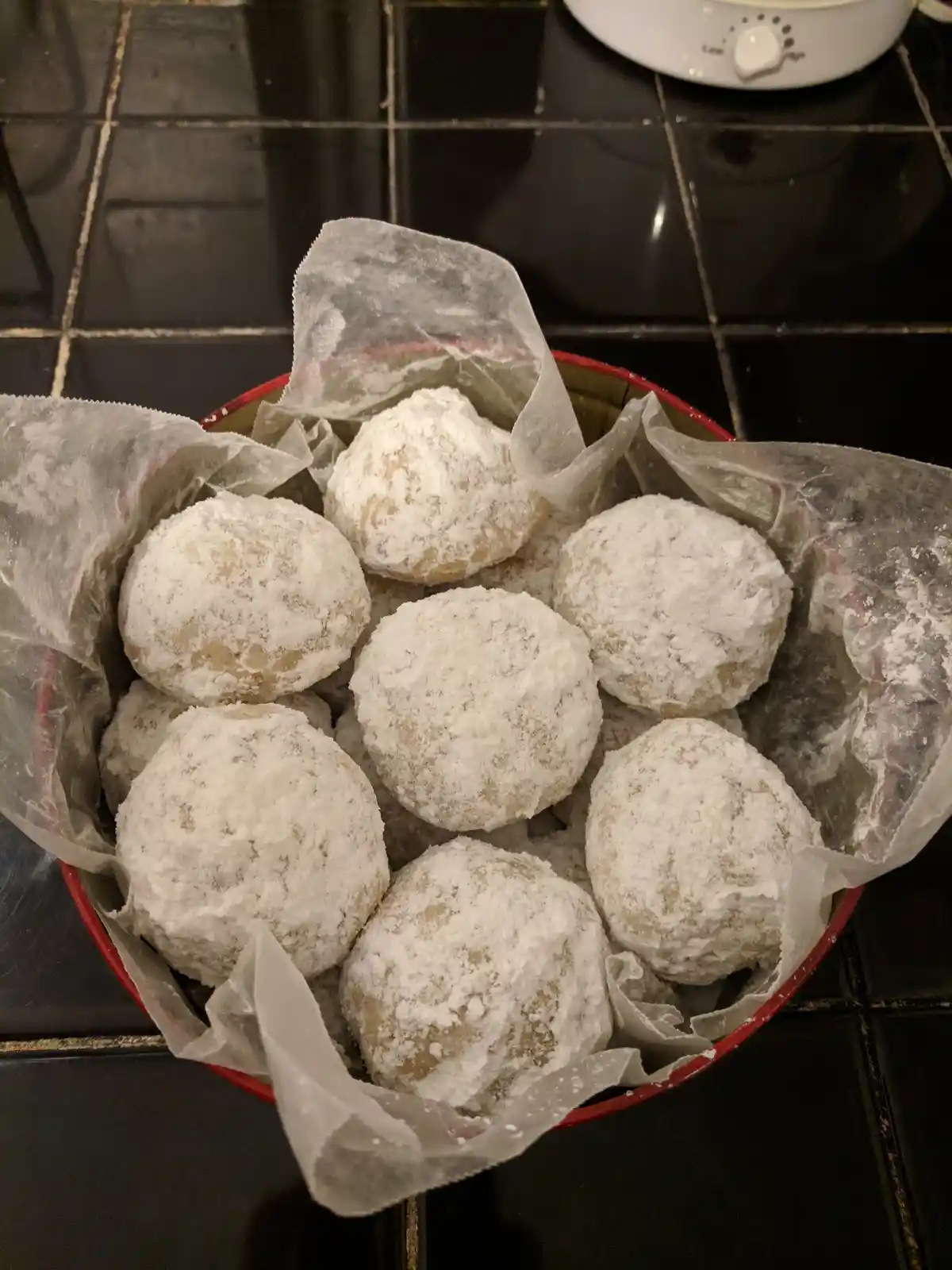 A red bowl lined with parchment paper sits on a black tiled kitchen counter, filled with round, white-frosted cookies…