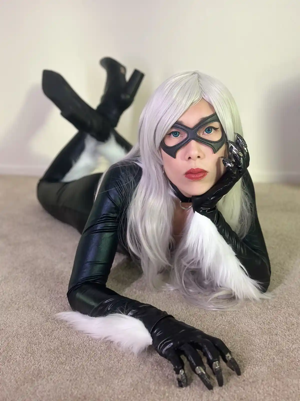 A person in a black and white cat-themed costume with long silver wig, black mask, and clawed gloves lies on a beige carpet…