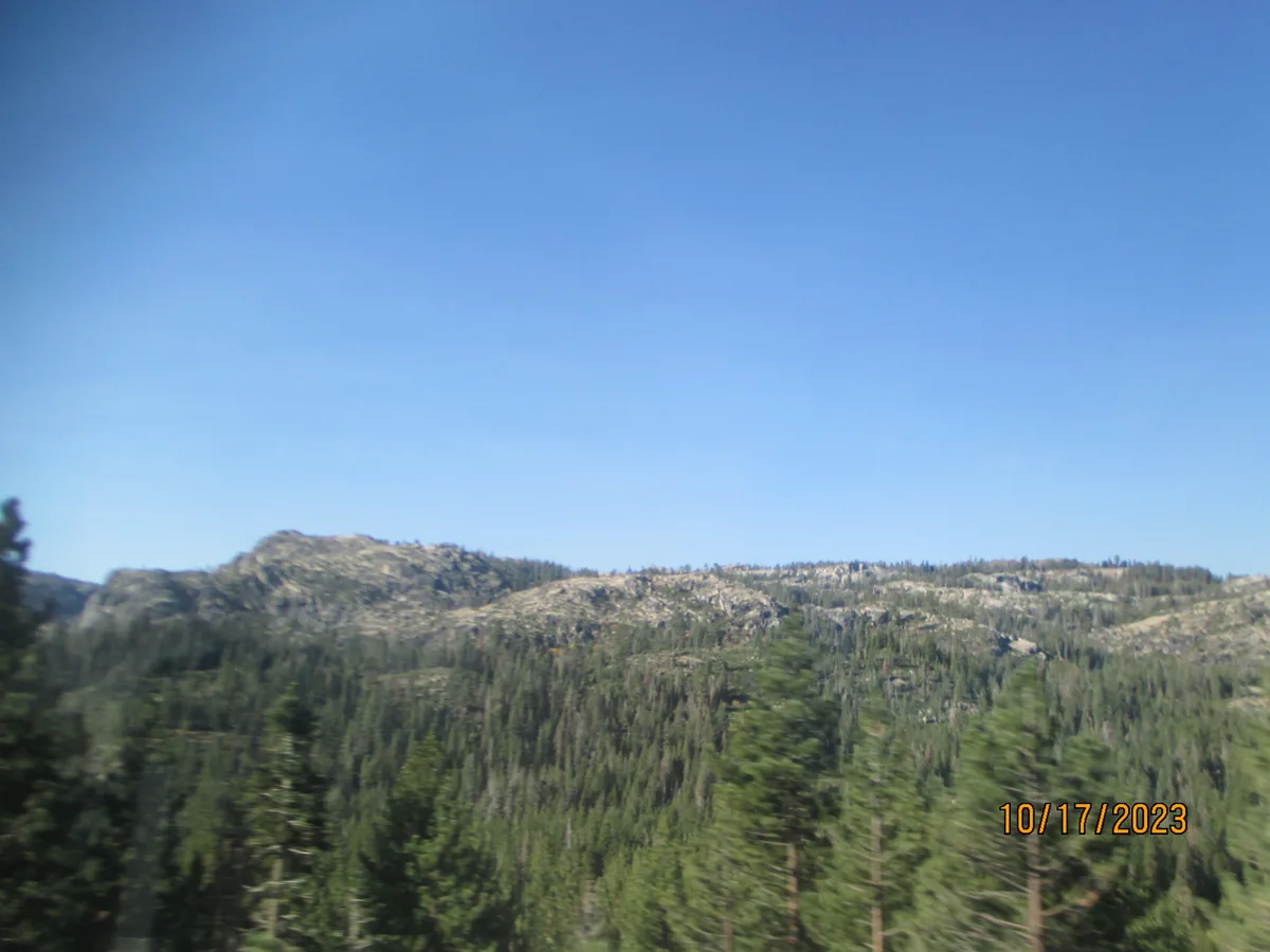 A blurry photograph of a forested mountain range under a clear blue sky, with a date stamp '10/17/2023' in orange text in…