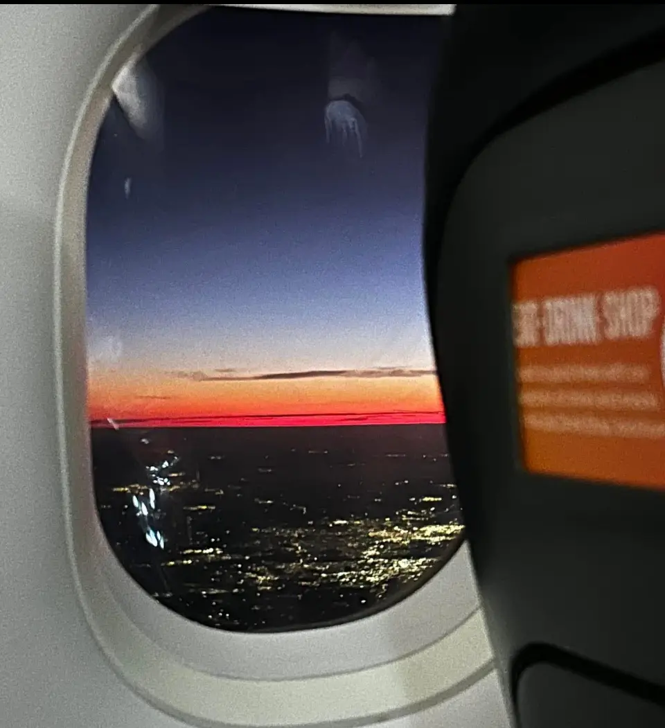 A view from an airplane window shows a stunning sunset over the ocean, with vibrant orange and red hues blending into a dark…