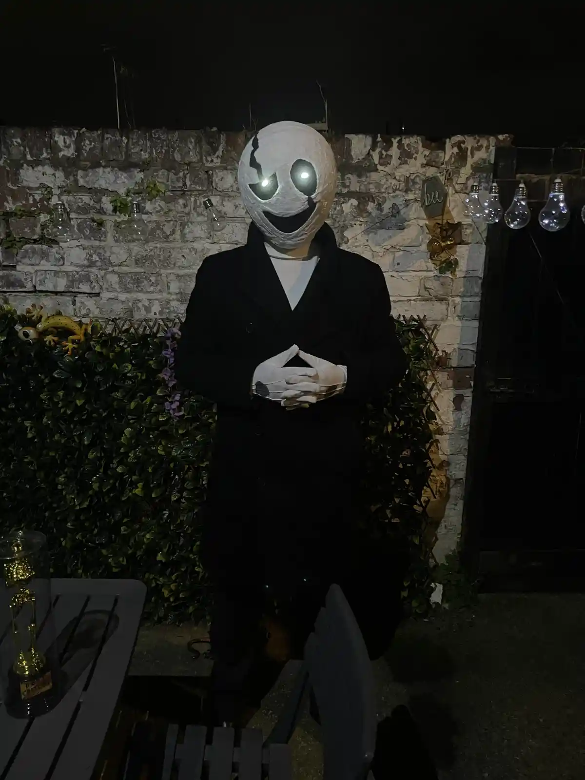 A person in a homemade Halloween costume stands at night, wearing a white paper-mâché mask with glowing eyes and a wide…