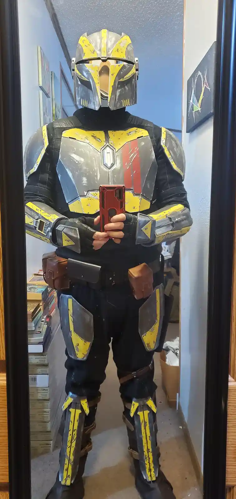A person in a full-body Mandalorian armor costume with yellow and gray weathered paint, standing in a hallway mirror and…