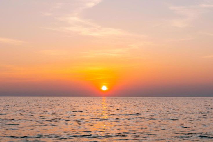 A serene sunset over calm water with the sun low on the horizon, casting orange and pink hues across the sky and reflecting…