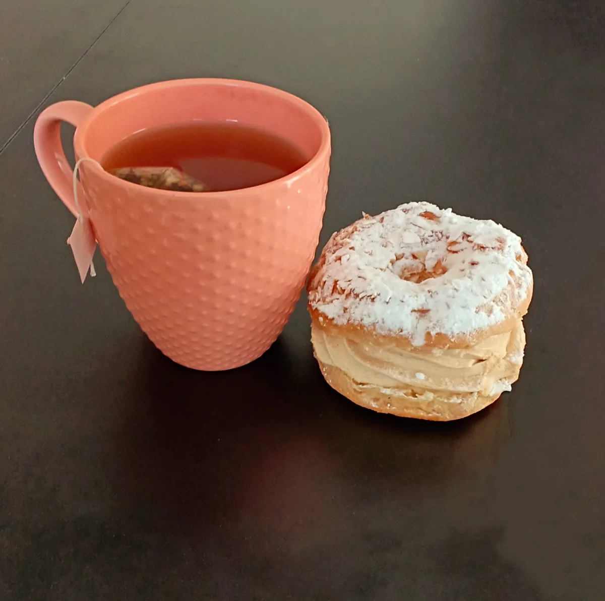 A peach-colored textured mug filled with tea and a tea bag tag hanging over the side sits next to a powdered sugar-dusted…