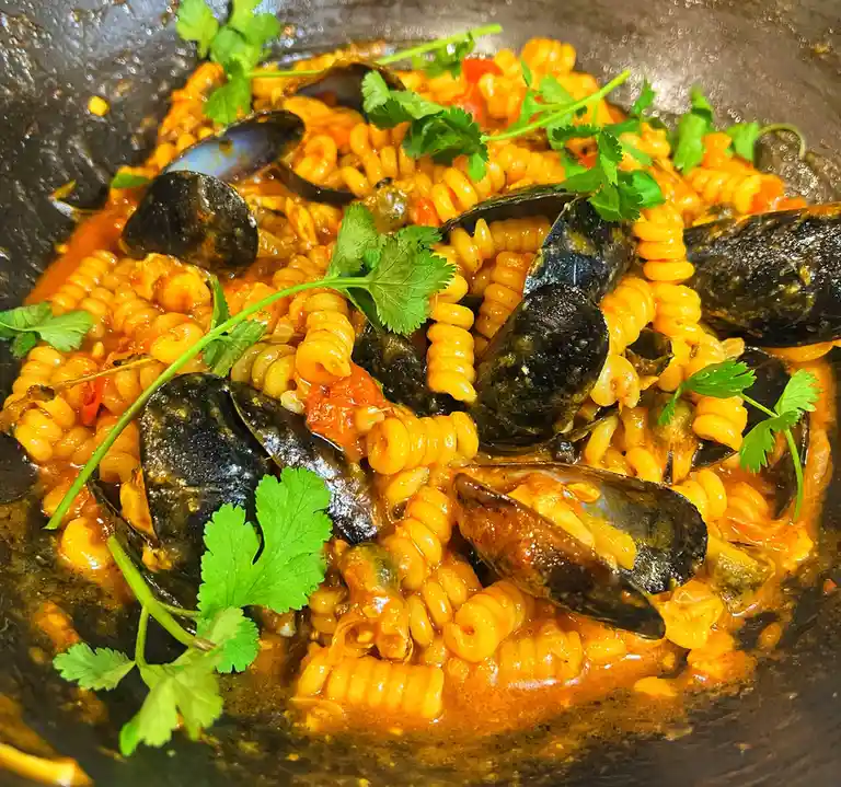 A close-up photo of a dish featuring spiral pasta in a red sauce, mixed with numerous open mussels and garnished with fresh…