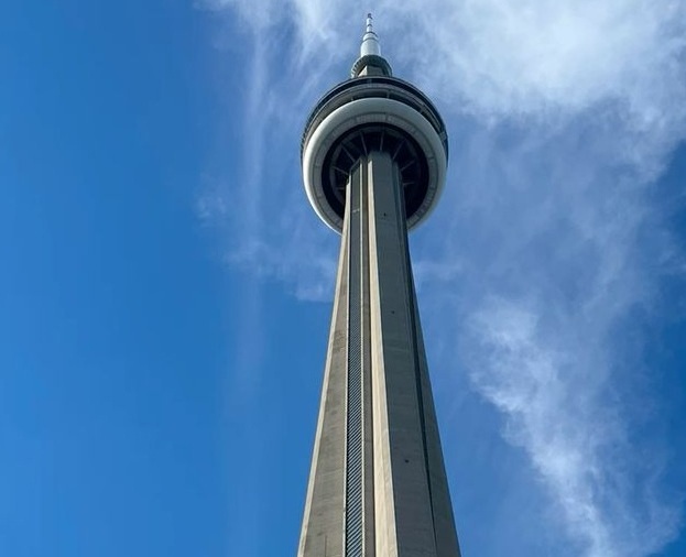 A low-angle photograph of the CN Tower in Toronto against a blue sky with wispy clouds. The image is overlaid with the text…
