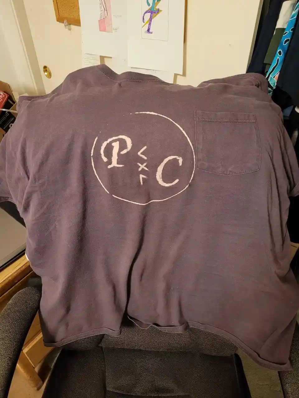 A dark gray t-shirt is draped over a chair, showing a hand-drawn white circle on the back with the letters 'P' and 'C'…