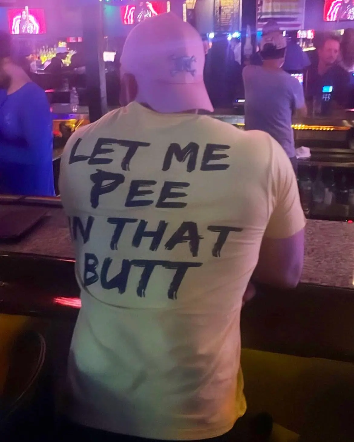A person viewed from behind is sitting at a bar, wearing a light-colored t-shirt with the phrase 'LET ME PEE IN THAT BUTT'…