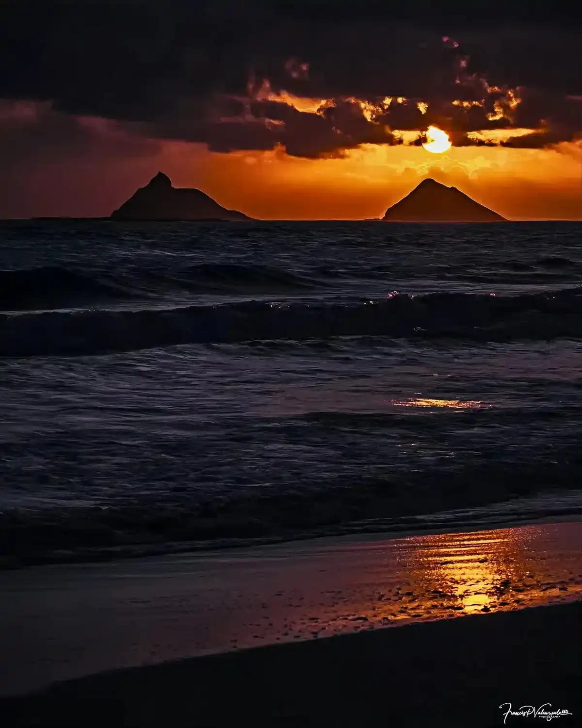 A dramatic sunset over a beach with two dark, pointed islands silhouetted against a fiery orange sky. The sun is partially…