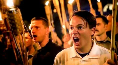 A man in a light-colored polo shirt is screaming with his mouth wide open while holding a torch, surrounded by other men…