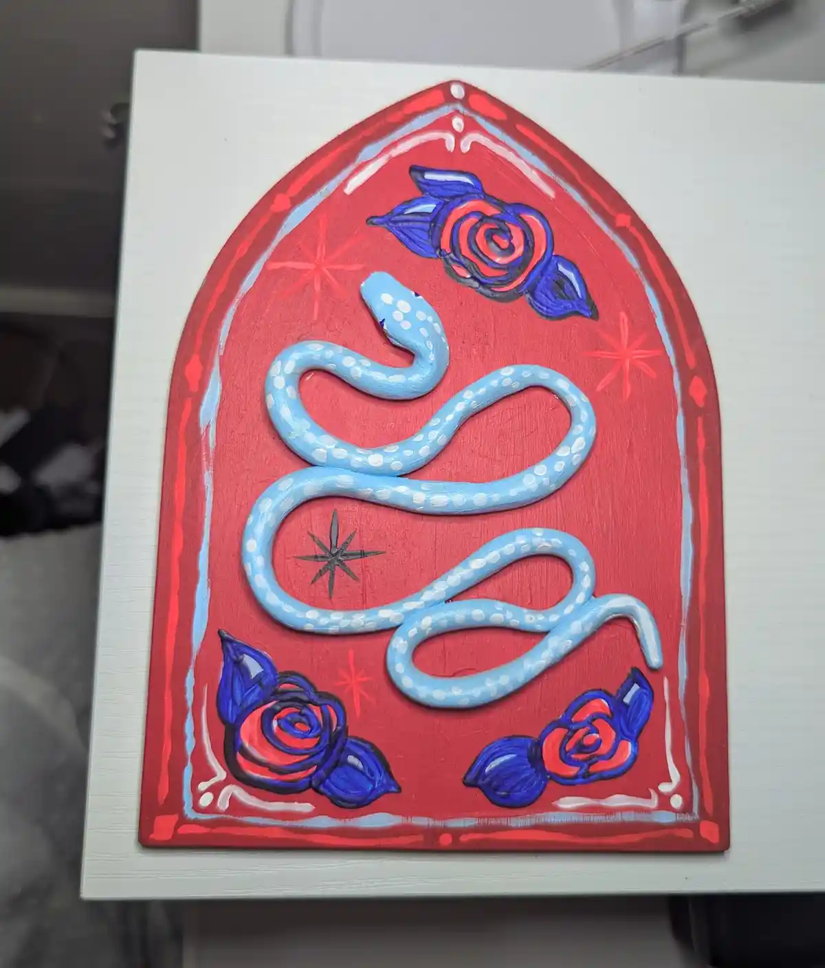 A hand-painted artwork on a white board depicts a light blue snake with white spots coiled within a red arched frame. The…