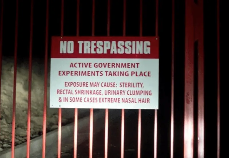 A red and white 'NO TRESPASSING' sign is mounted on a red metal fence at night. The sign warns of 'ACTIVE GOVERNMENT…