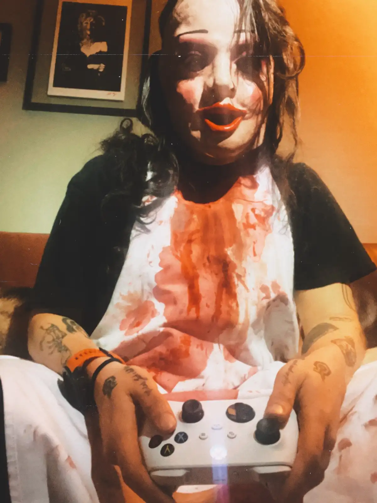 A person wearing a scary, painted mask with bright red lips and smeared orange-red stains on their white t-shirt and arms…