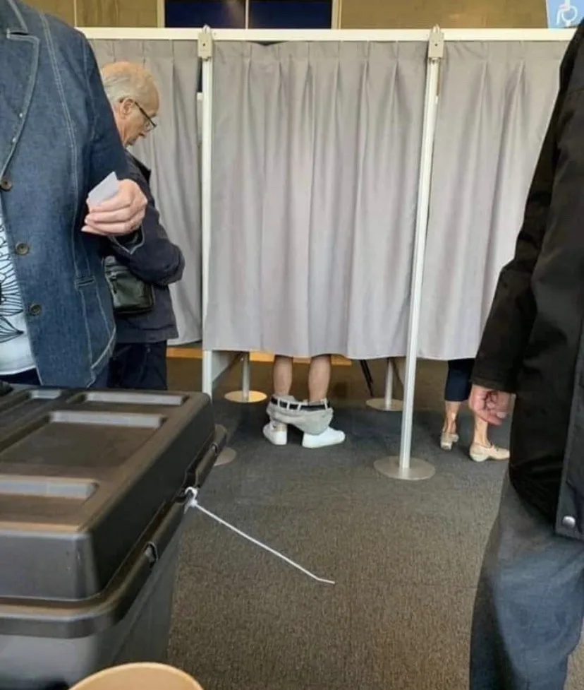 A candid photo taken inside a voting booth area shows a person standing behind a privacy curtain, visible only from the…