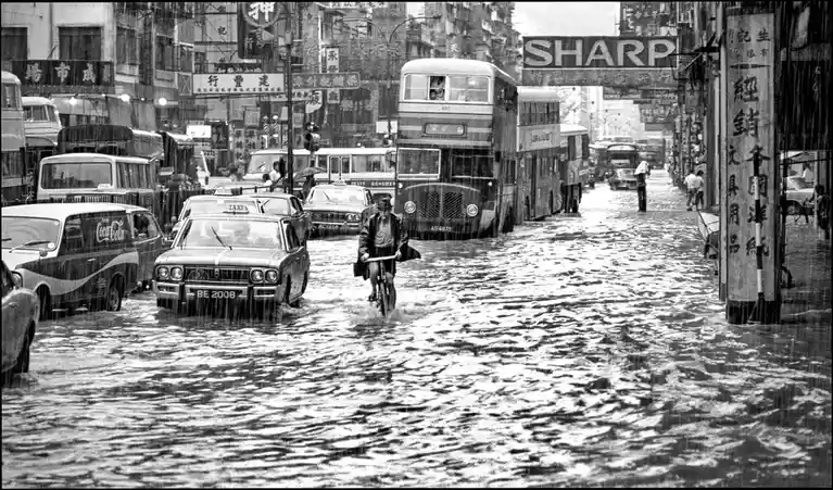 A black-and-white photo shows a flooded city street with vintage cars, double-decker buses, and a man riding a bicycle…