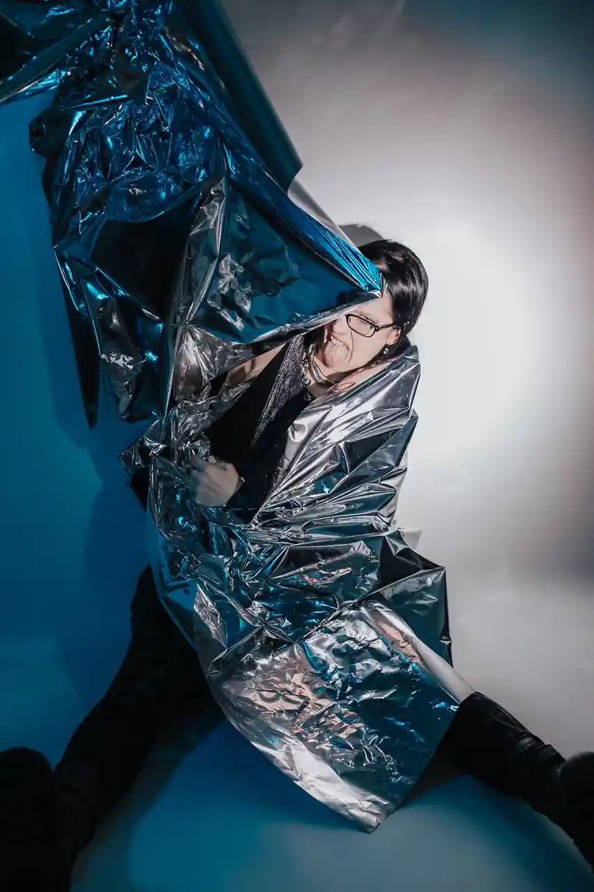 A person with dark hair and glasses is sitting on the floor, wrapped in a crinkled, reflective blue thermal blanket. They…