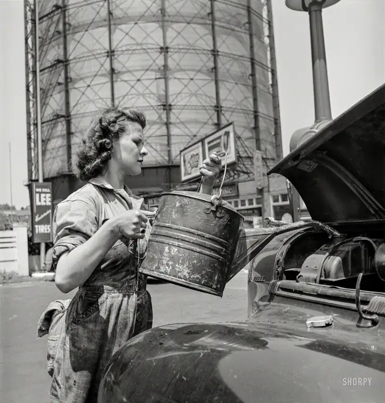 a black-and-white photo from the 1940s shows a woman in overalls pouring liquid from a metal bucket into the engine of a car…