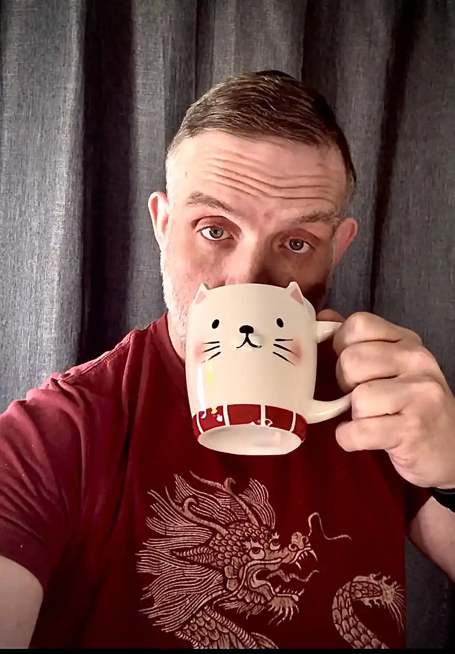 A man with a beard and short hair, wearing a red t-shirt with a dragon design, holds a white mug with a cartoon cat face…