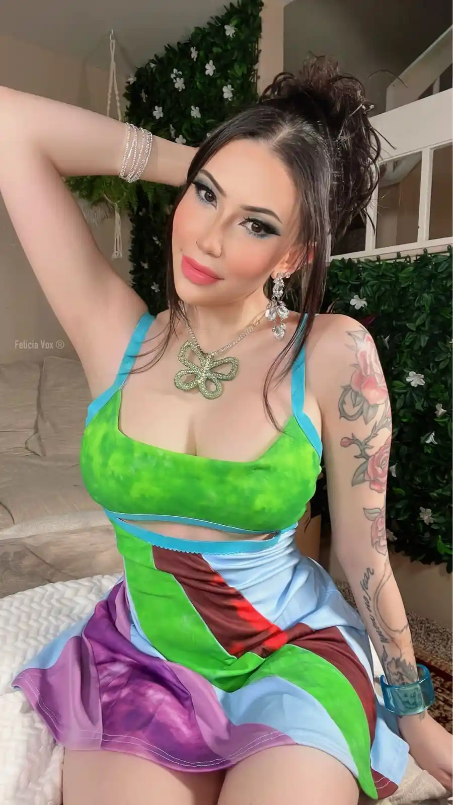 A woman with dark hair in a messy bun and heavy makeup poses in a colorful, multi-toned dress with green, purple, blue, and…