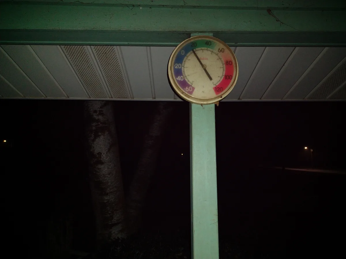 A close-up of an outdoor thermometer mounted on a light green pole under a covered structure at night. The thermometer's…