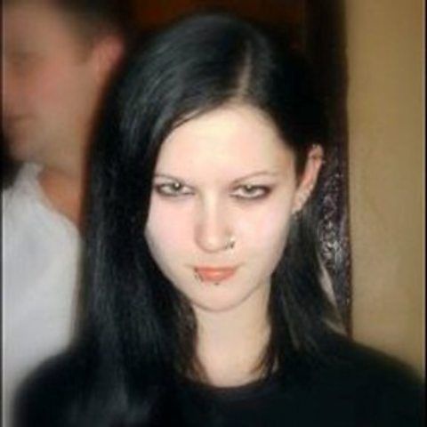 A woman with long black hair, dark eye makeup, and a lip piercing stares directly at the camera with a neutral expression…