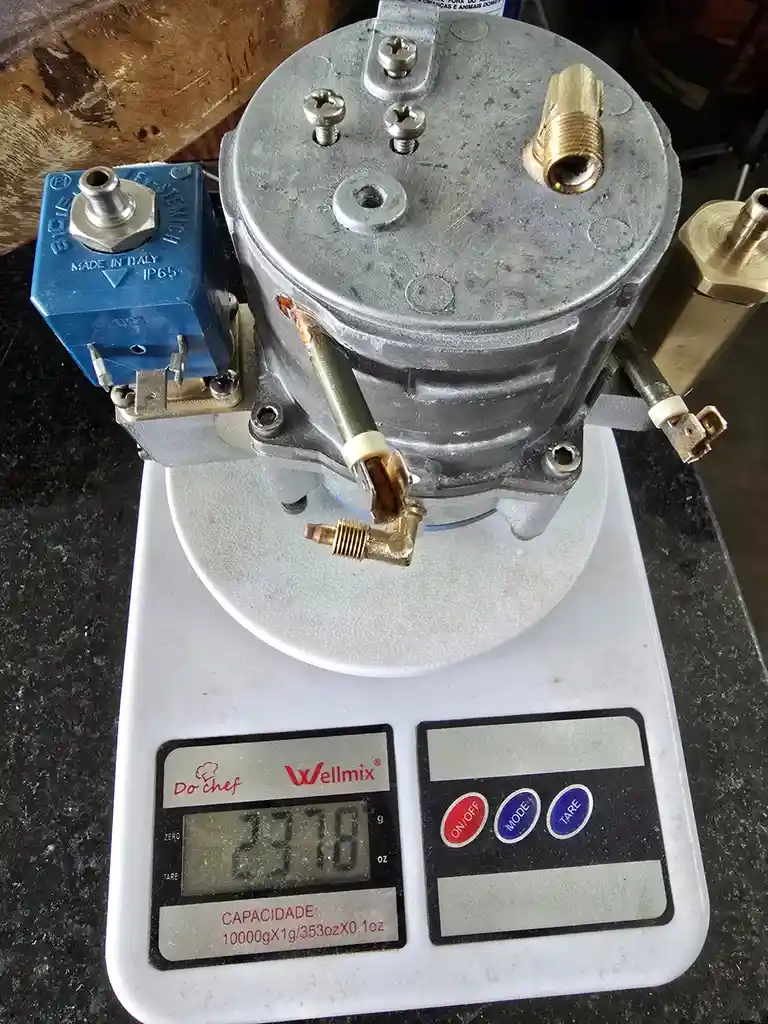 A silver industrial-looking pump or compressor with brass fittings sits on a white digital kitchen scale. The scale's…