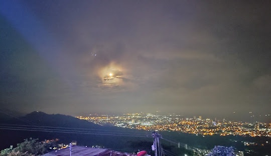 A night sky photograph showing a bright moon partially obscured by wispy clouds above a sprawling cityscape illuminated by…