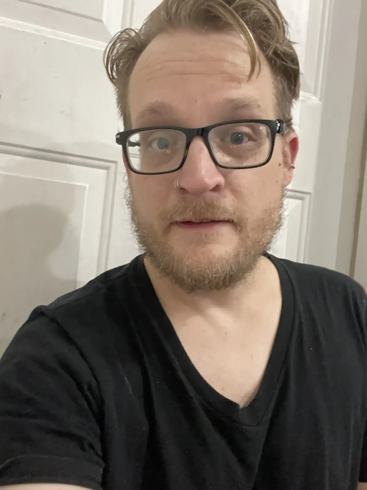 A close-up selfie of a man with light brown hair, a beard, and black-rimmed glasses, wearing a black v-neck t-shirt…